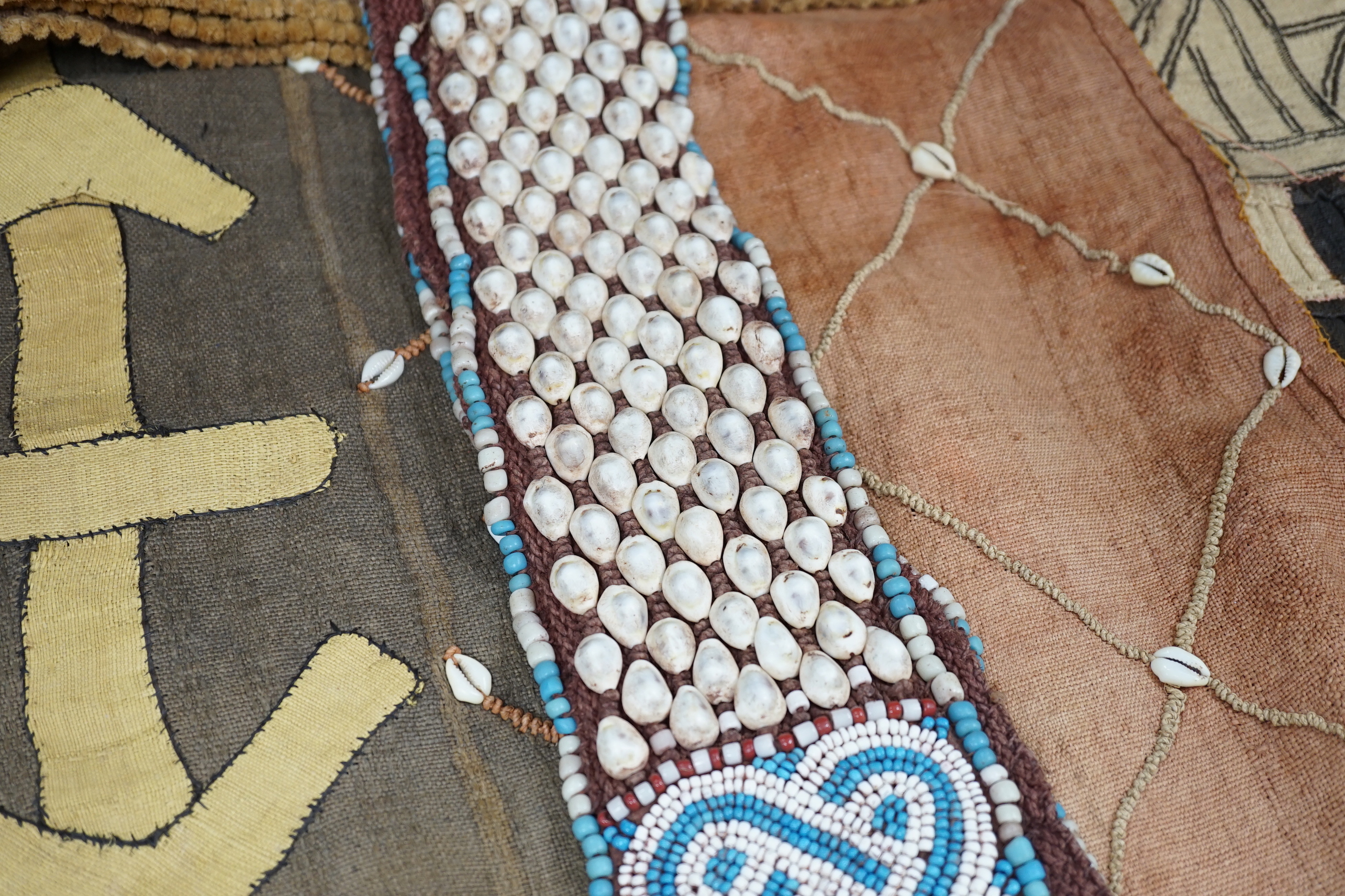 Three African tribal hangings, embroidered with coloured hand dyed threads and appliquéd shells on smaller handwoven cloth panels all sewn together to form the hangings, including a chieftains coloured bead worked and sh